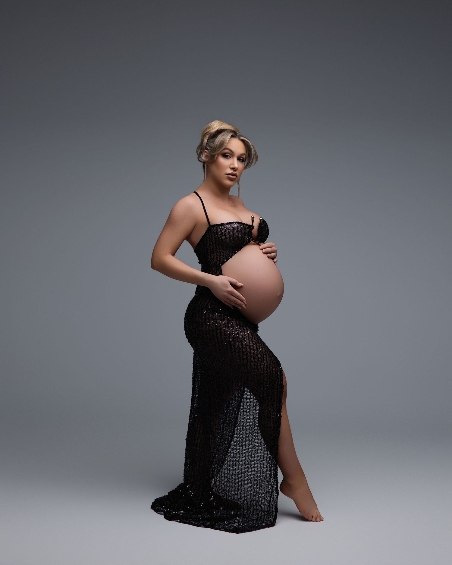 Person in a black sheer gown poses elegantly for a dramatic maternity photo against a gray studio backdrop.