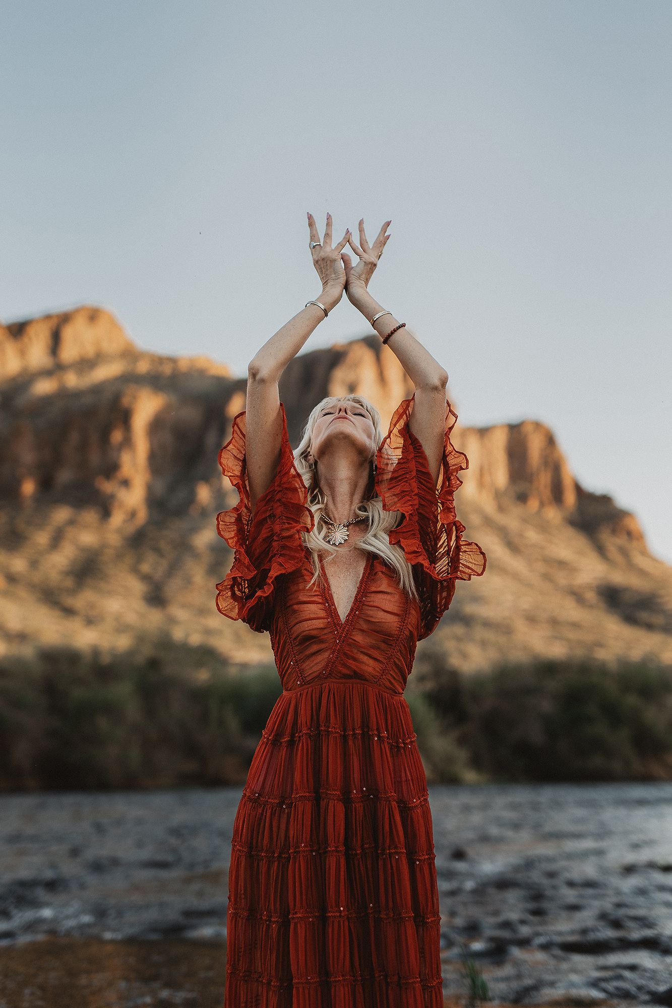 A person in a flowing red dress strikes an expressive pose against a mountainous desert landscape at sunset.