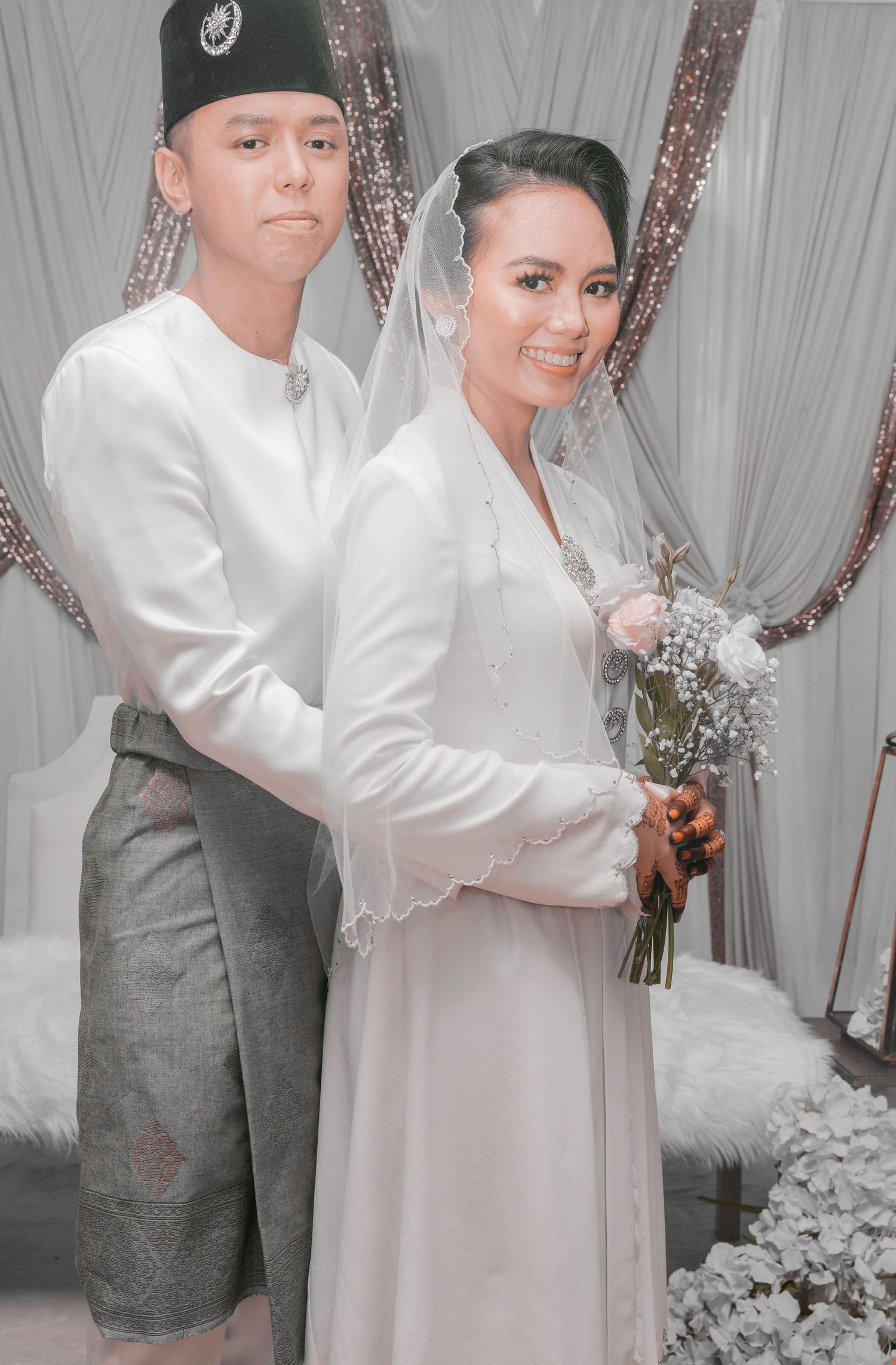 A couple in traditional Vietnamese wedding attire pose together against a silver backdrop with ornate decorations.