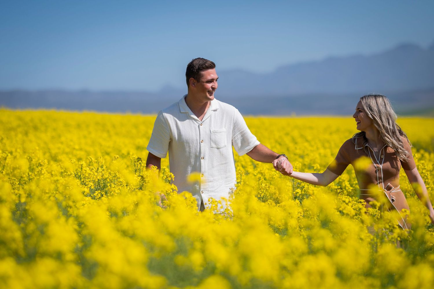 A couple holds hands while walking through a vibrant yellow canola field with mountains in the background.