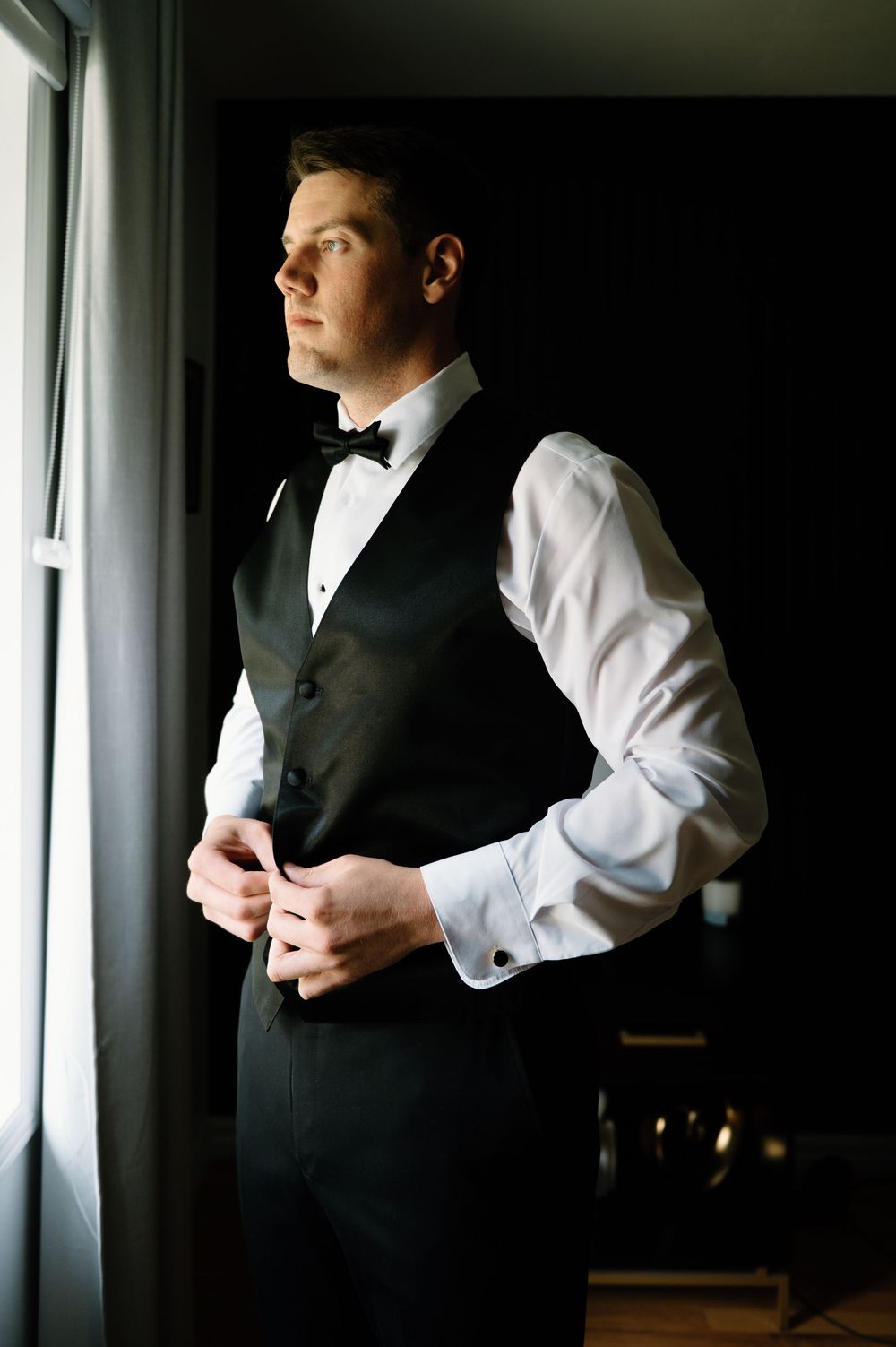 Figure in formal black vest and white shirt adjusts clothing near window.