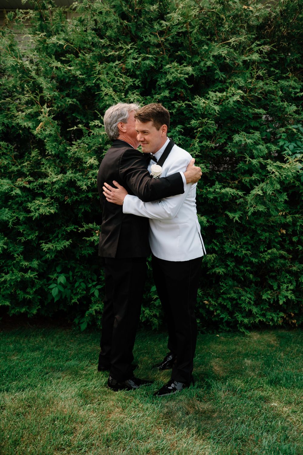 A couple in formal attire dances playfully on grass with green bushes in background.