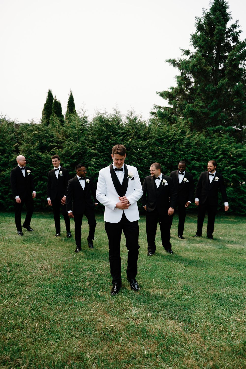 Wedding party members in black suits and white jacket stand in formation on grass.