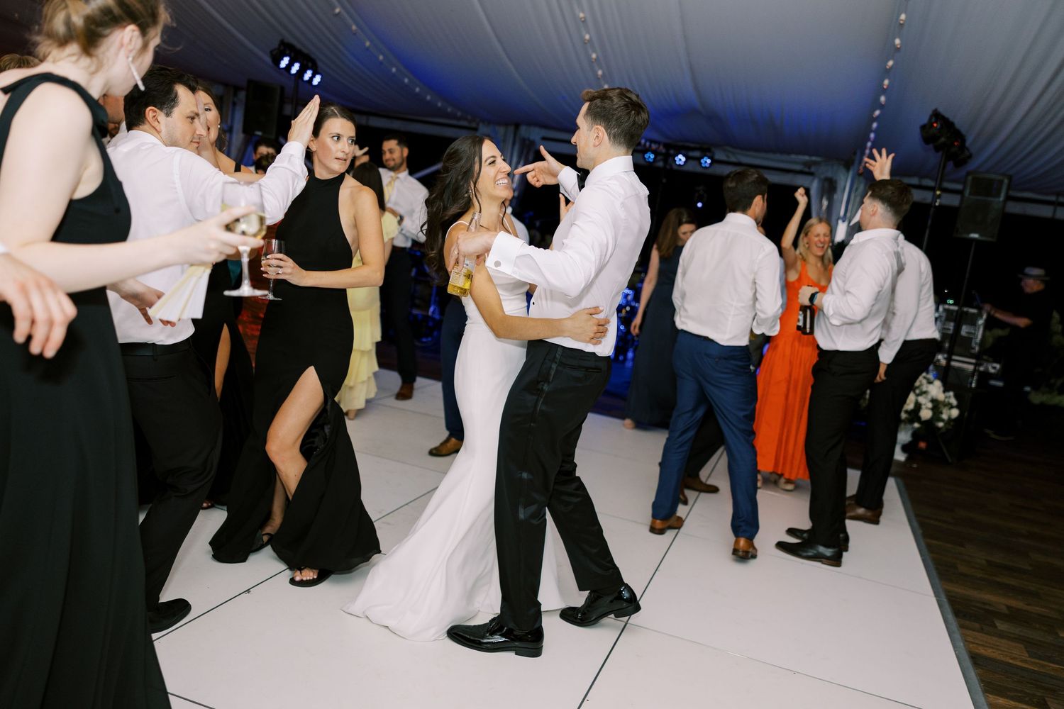 A group of guests dancing during an evening wedding reception on a white dance floor under blue lighting.