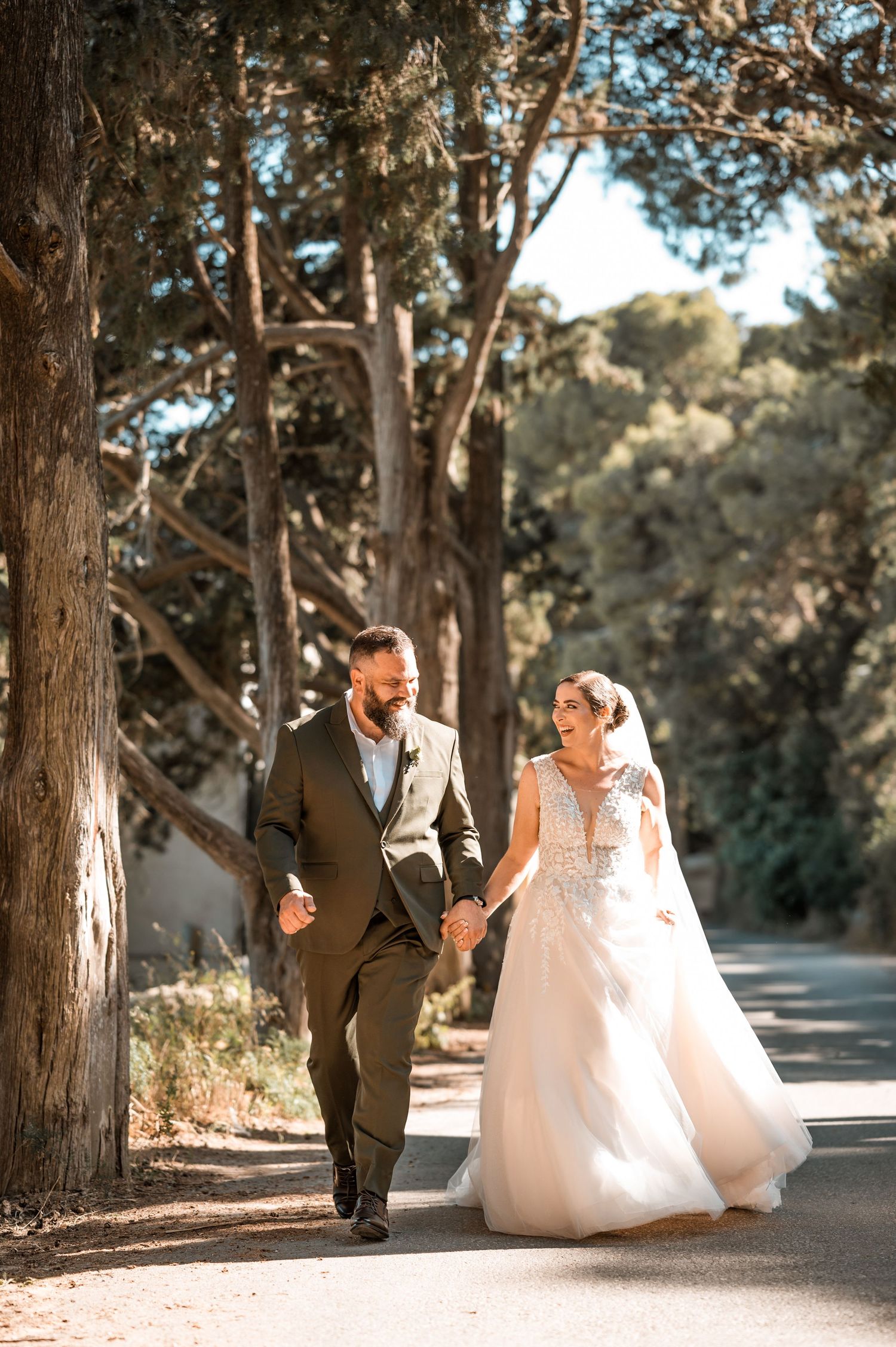 Wedding couple walking through a tree-lined path in formal attire.