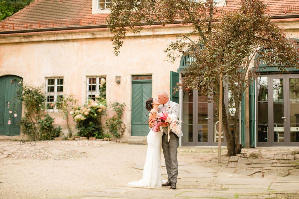 A couple embraces outside a charming rustic building with pink walls and green doors in a romantic courtyard setting.