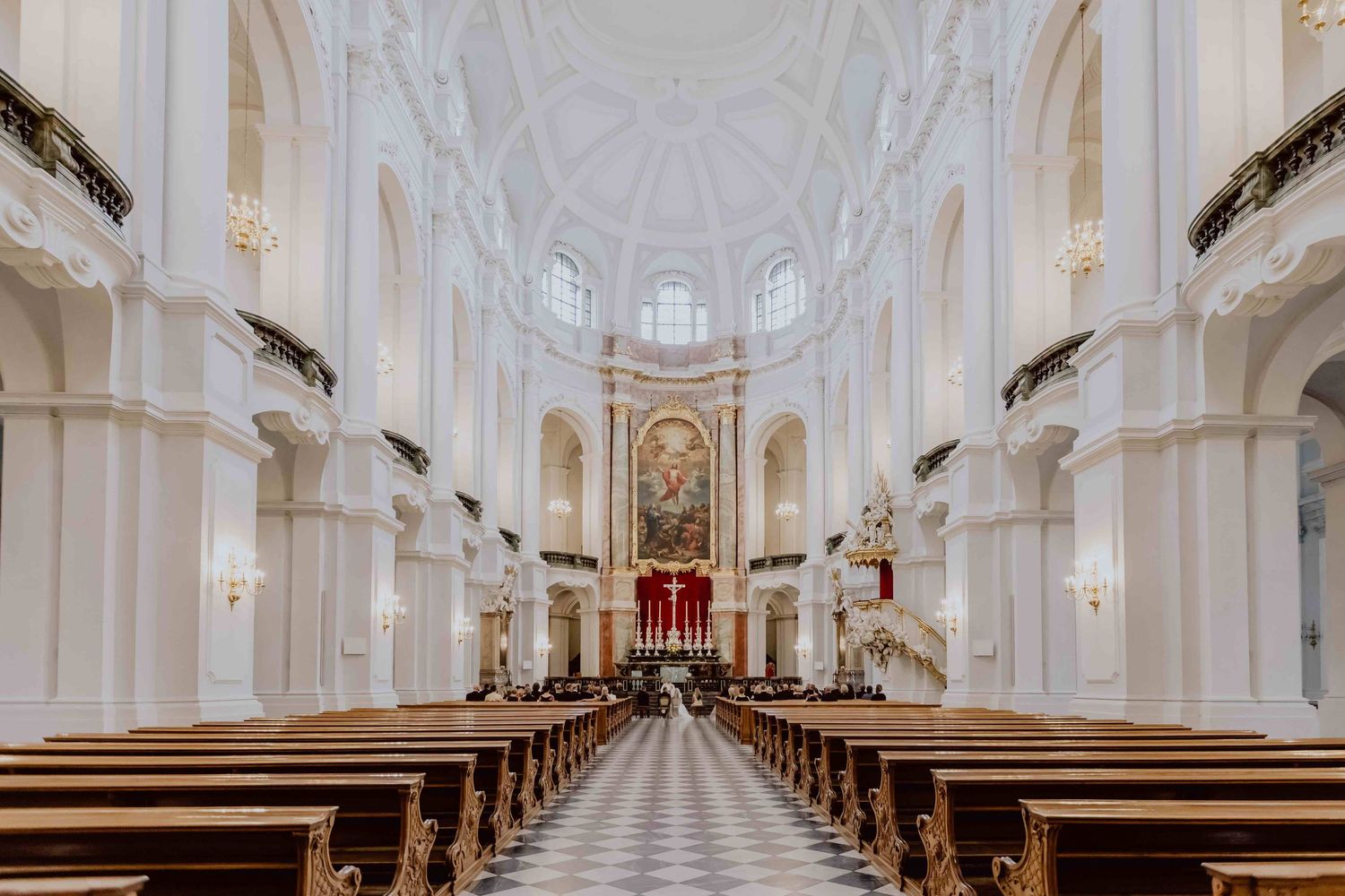 Interior view of an ornate white baroque church, Hofkirche in Dresden, with symmetrical pews leading to a grand altar and domed ceiling.