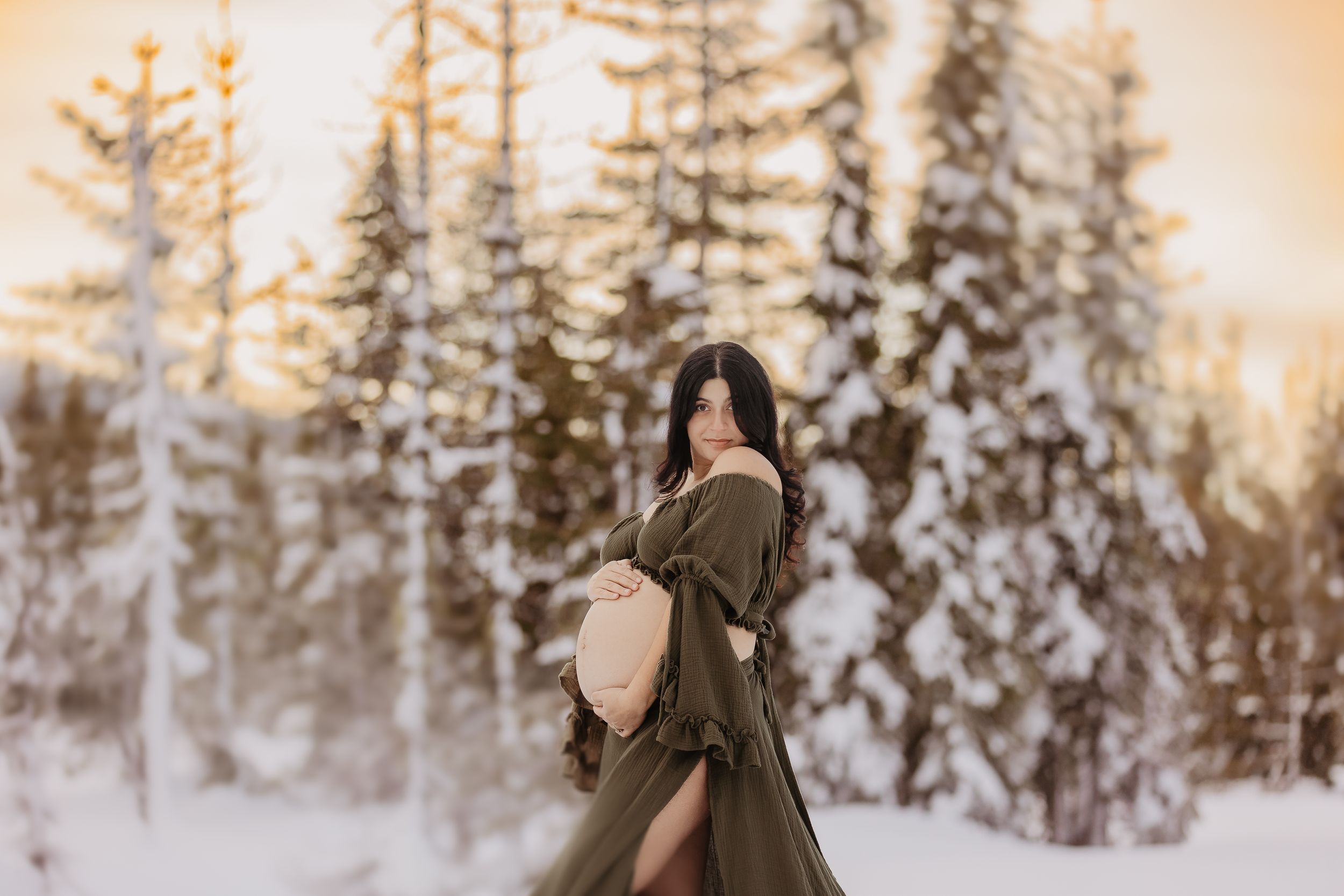 Winter maternity photos in snowy forest with sun glowing through snow-covered trees.