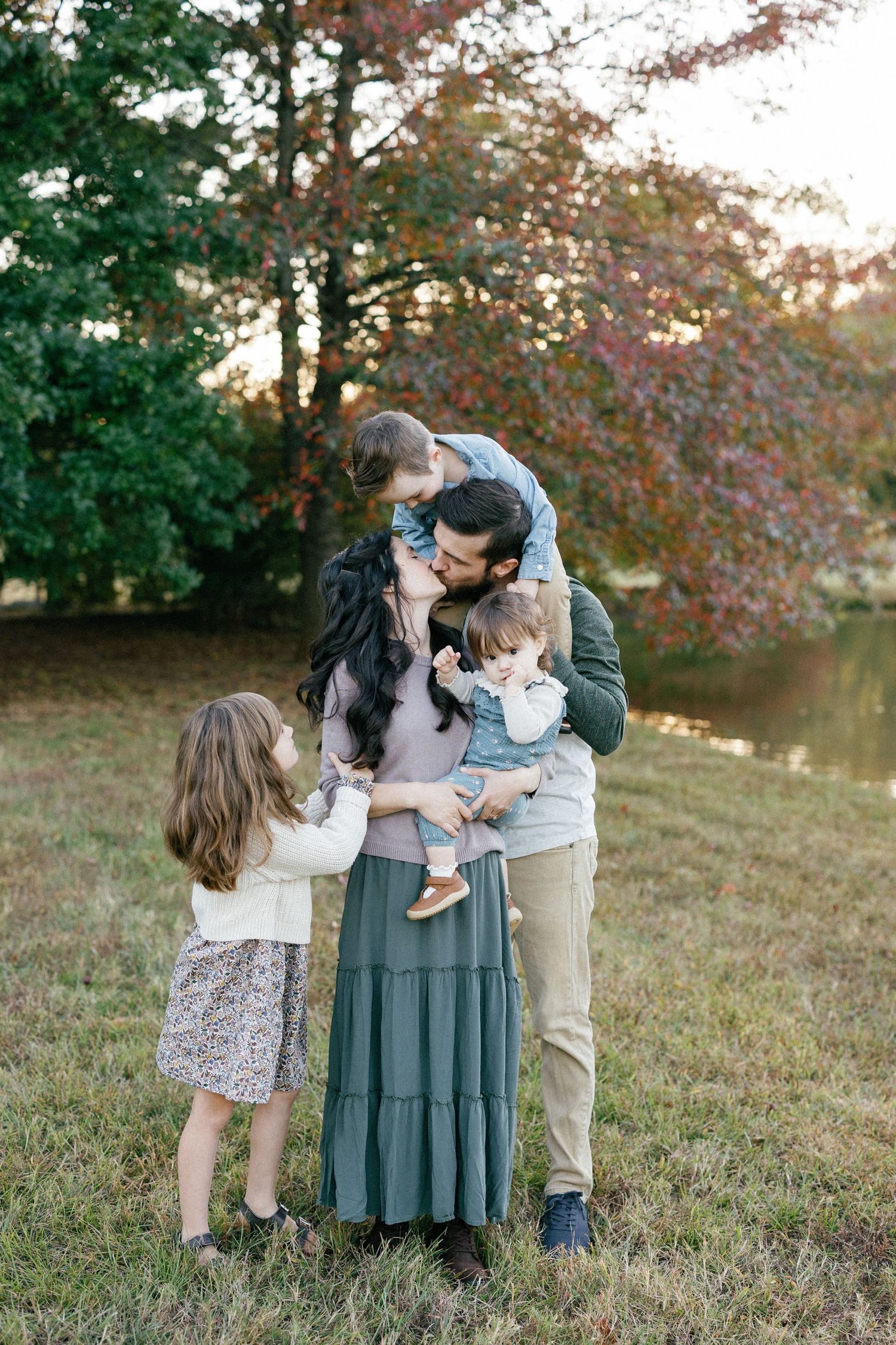 Expecting parents in white and khaki outfits pose with an ultrasound in a grassy outdoor setting at sunset.