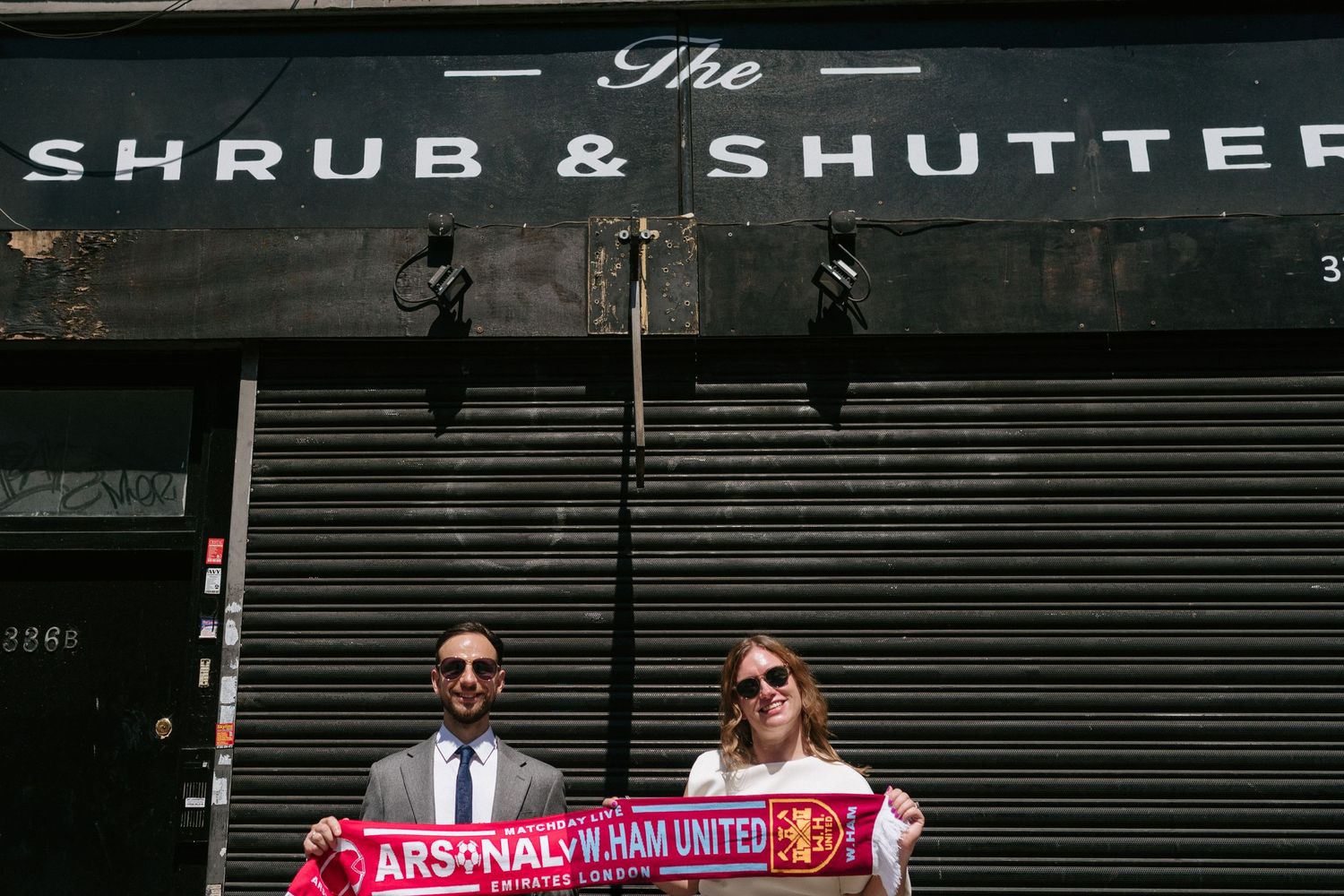 Two figures hold up a red soccer scarf in front of a closed storefront with text reading 'The Shrub & Shutter'.