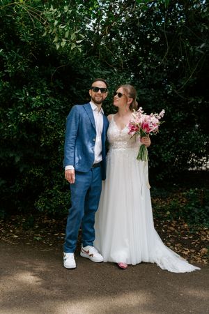 A couple poses outdoors on their wedding day, with the bride holding a pink bouquet against lush green foliage.