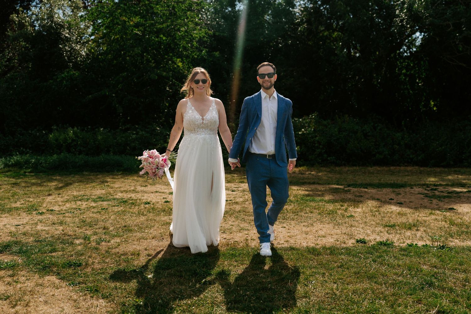 A couple walking together on grass in wedding attire, with the bride holding a pink bouquet in a sunny outdoor setting.