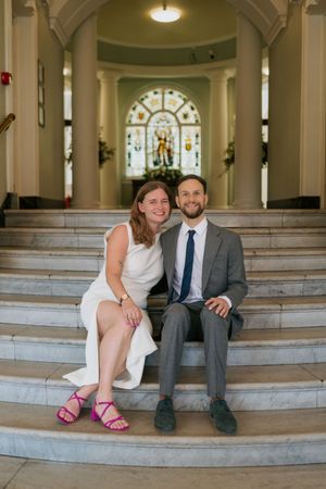 A couple sits on grand marble steps inside a building with ornate architectural details and stained glass.
