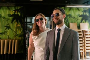 A cheerful pair in sunglasses smile together, with one wearing a grey suit and tie and the other in a light colored top.
