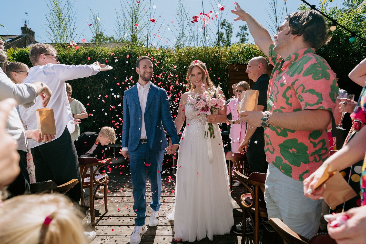 Happy newlyweds walk down the aisle during an outdoor garden wedding ceremony as guests throw flower petals in celebration.