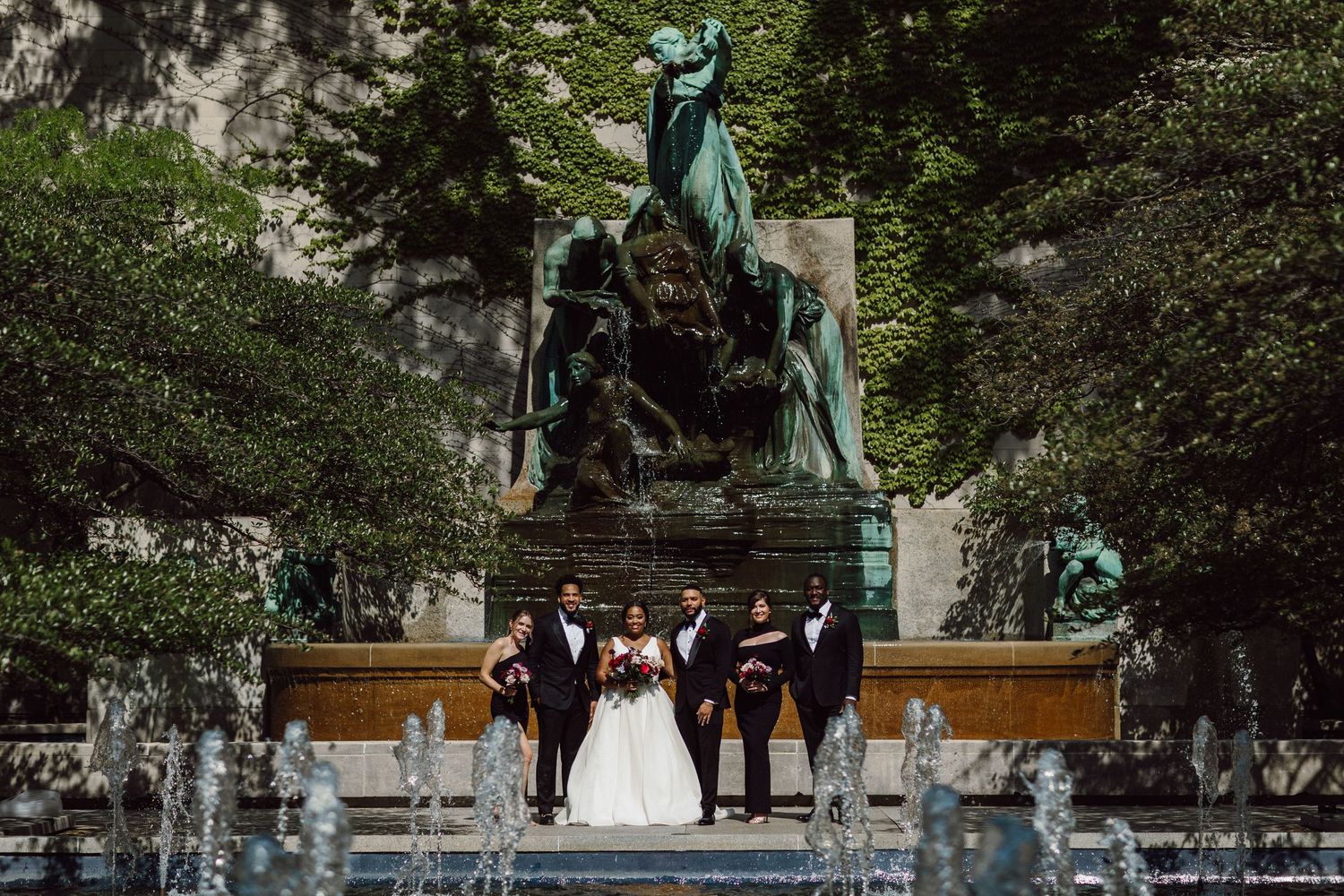 Couple poses near outdoor fountain with green statues in background.