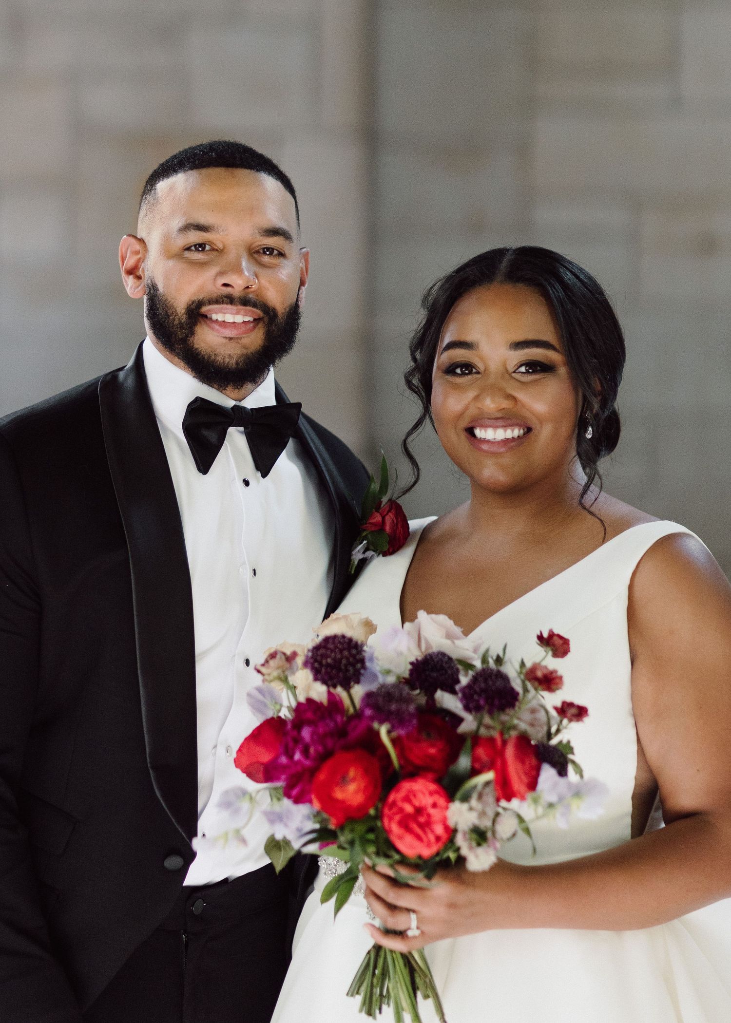Newlyweds share intimate moments while holding vibrant red and purple bouquet.
