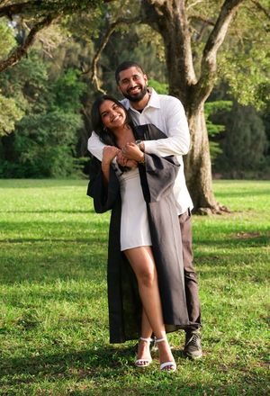 Couple embraces and smiles together outdoors on green grass with large oak trees behind them.