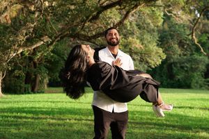 Man lifts and spins smiling graduate in black gown outdoors on green lawn under large tree.