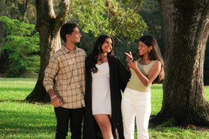 Graduate in white dress and gown laughs with two friends under oak trees on green lawn.