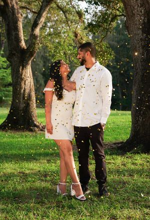 Couple stands together smiling as golden confetti falls around them on a sunny green lawn.