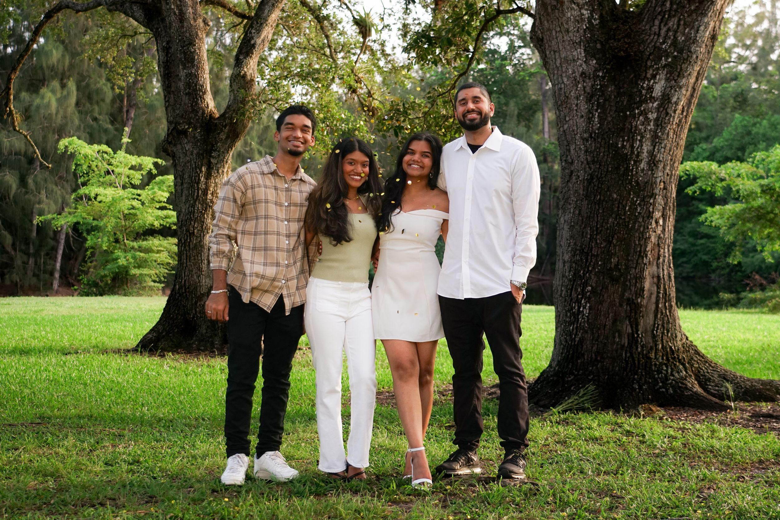 Family of four smiling together outdoors under large oak trees on green grass.