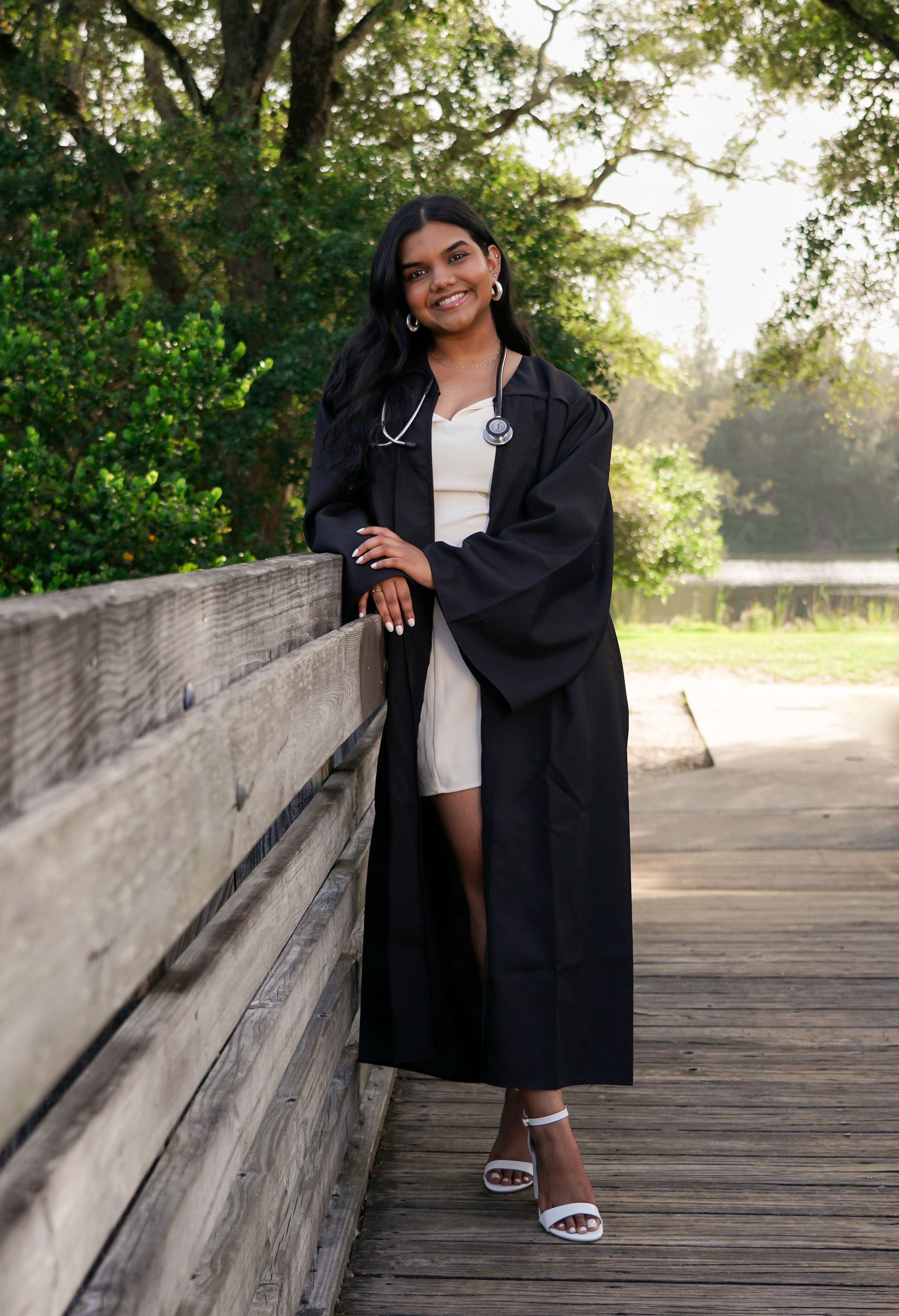 Young woman in black graduation gown with stethoscope leans on wooden bridge railing outdoors.