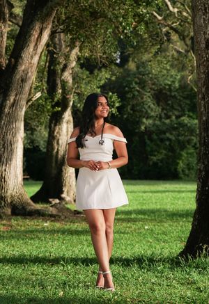 Young woman in white off-shoulder dress stands between trees on sunny green lawn smiling.