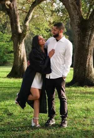 Graduate in black gown and man in white shirt share a loving glance under trees in park.