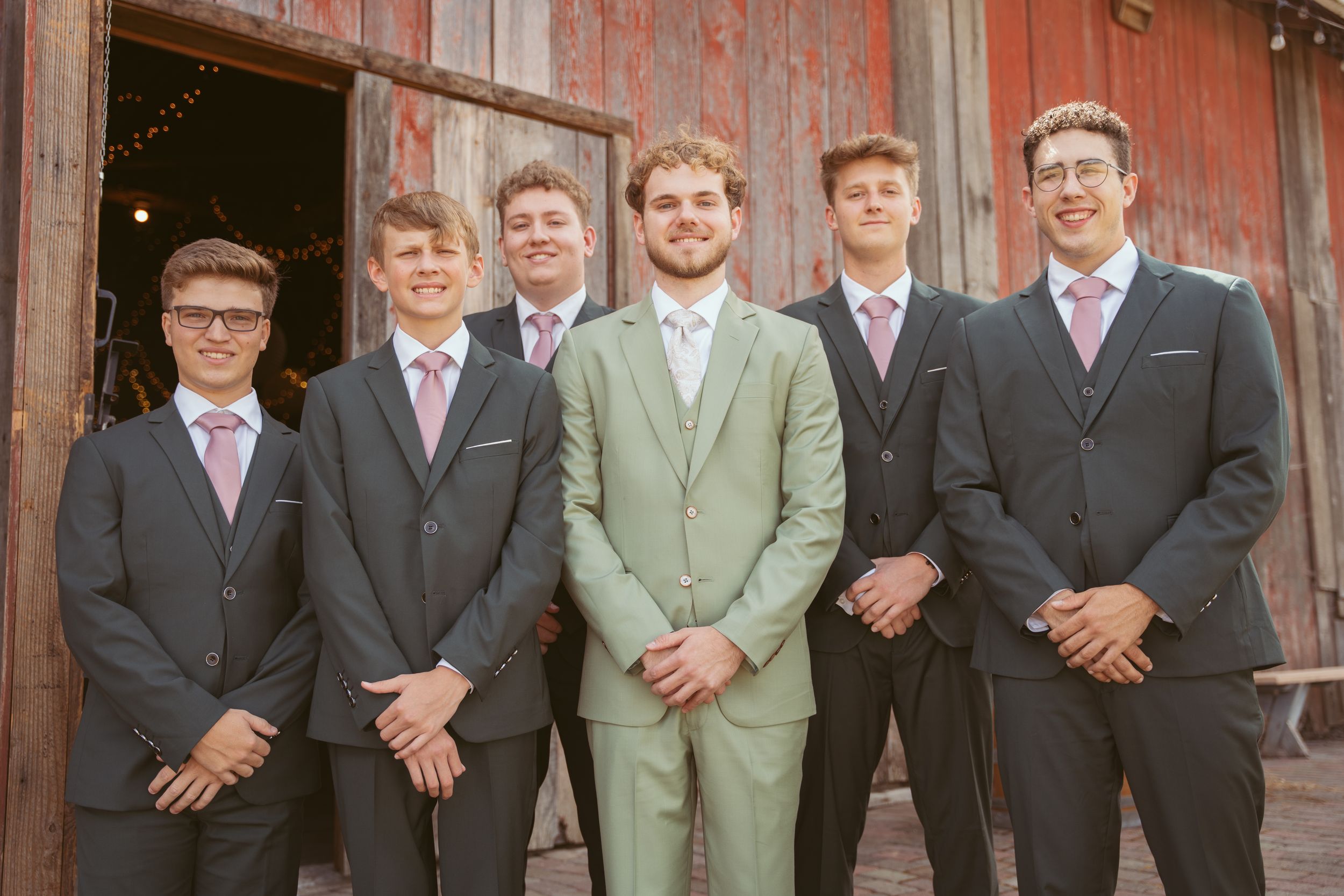 A groom in light green suit stands with groomsmen wearing dark suits in front of a rustic red barn wall.