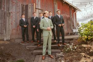 A wedding party in formal attire stands outside a rustic barn with string lights, featuring groomsmen in black and sage green suits.