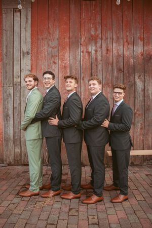 Group of groomsmen in formal suits line up playfully against rustic red barn wall at wedding venue.