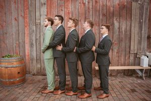 Five groomsmen in matching suits stand in line against rustic wooden barn wall with brown barrel decor.