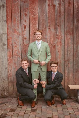 Four sequential photos show groomsmen in green and black suits posing playfully on rustic brick patio against wooden wall.