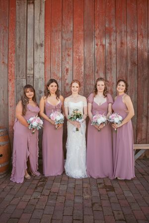 Wedding party in mauve bridesmaid dresses and white bridal gown pose against rustic red barn wall.