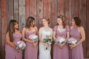 A bridal party wearing mauve bridesmaid dresses holds pink and white bouquets while posing against a rustic wooden wall.