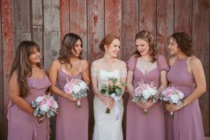 A bridal party wearing dusty rose colored dresses poses with white and pink bouquets against a rustic wooden wall.