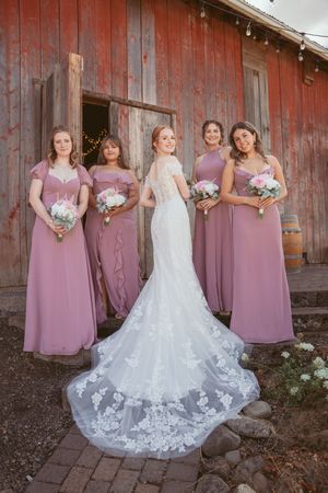 A bridal party in mauve bridesmaid dresses poses against a rustic red barn with a stunning white lace wedding gown.