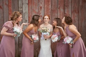 A bridal party in dusty rose dresses poses together with white and pink bouquets against a rustic wooden barn wall.
