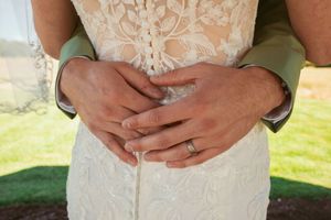 Close up of hands wearing wedding rings against white lace wedding dress and sage green suit.