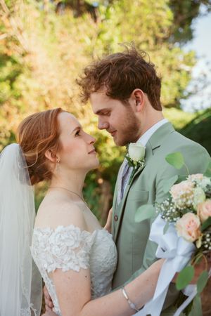 Romantic wedding moment outdoors with green suit and white lace dress against natural scenery.