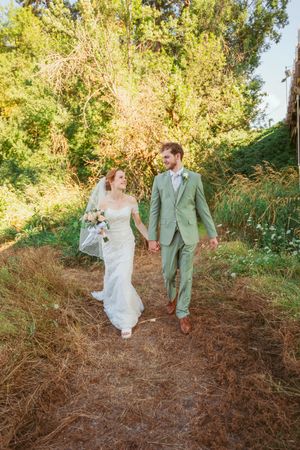 Couple in wedding attire walk hand in hand through scenic golden field with trees in background.