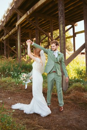 A couple celebrates joyfully under a rustic wooden structure at sunset, with the bride in white and groom in sage green.