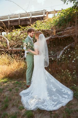A romantic outdoor wedding photo with a stunning lace train dress and sage green suit against a rustic bridge backdrop.