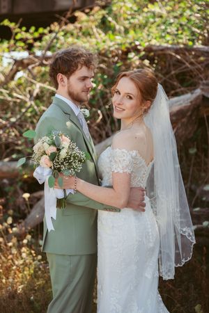 A wedding couple poses outdoors with the bride in lace and groom in sage green suit holding a pastel bouquet.