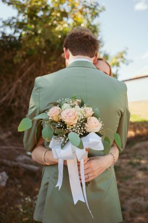 Wedding party members in sage green suits embrace while showing decorative floral boutonnieres during outdoor photoshoot.