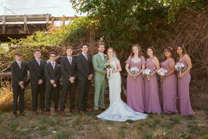 A wedding party in formal attire poses outdoors with bridesmaids in purple dresses and groomsmen in dark suits.