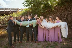 Wedding party in formal attire playfully posing outdoors with groomsmen in suits and bridesmaids in pink dresses.
