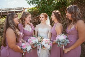 Wedding party in dusty rose bridesmaid dresses gather outdoors holding pink and white floral bouquets.