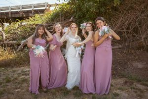 A group in matching mauve bridesmaid dresses and white wedding gown pose together while laughing outdoors.